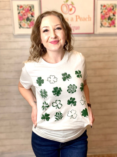 Model wearing our green clovers tee. The tee is short sleeve with clover sketches of different styles. The clovers are green in color and are in a 4 by 4 table form.