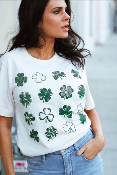 Model wearing our green clovers tee. The tee is short sleeve with clover sketches of different styles. The clovers are green in color and are in a 4 by 4 table form. Model pairs the top with light denim jeans.